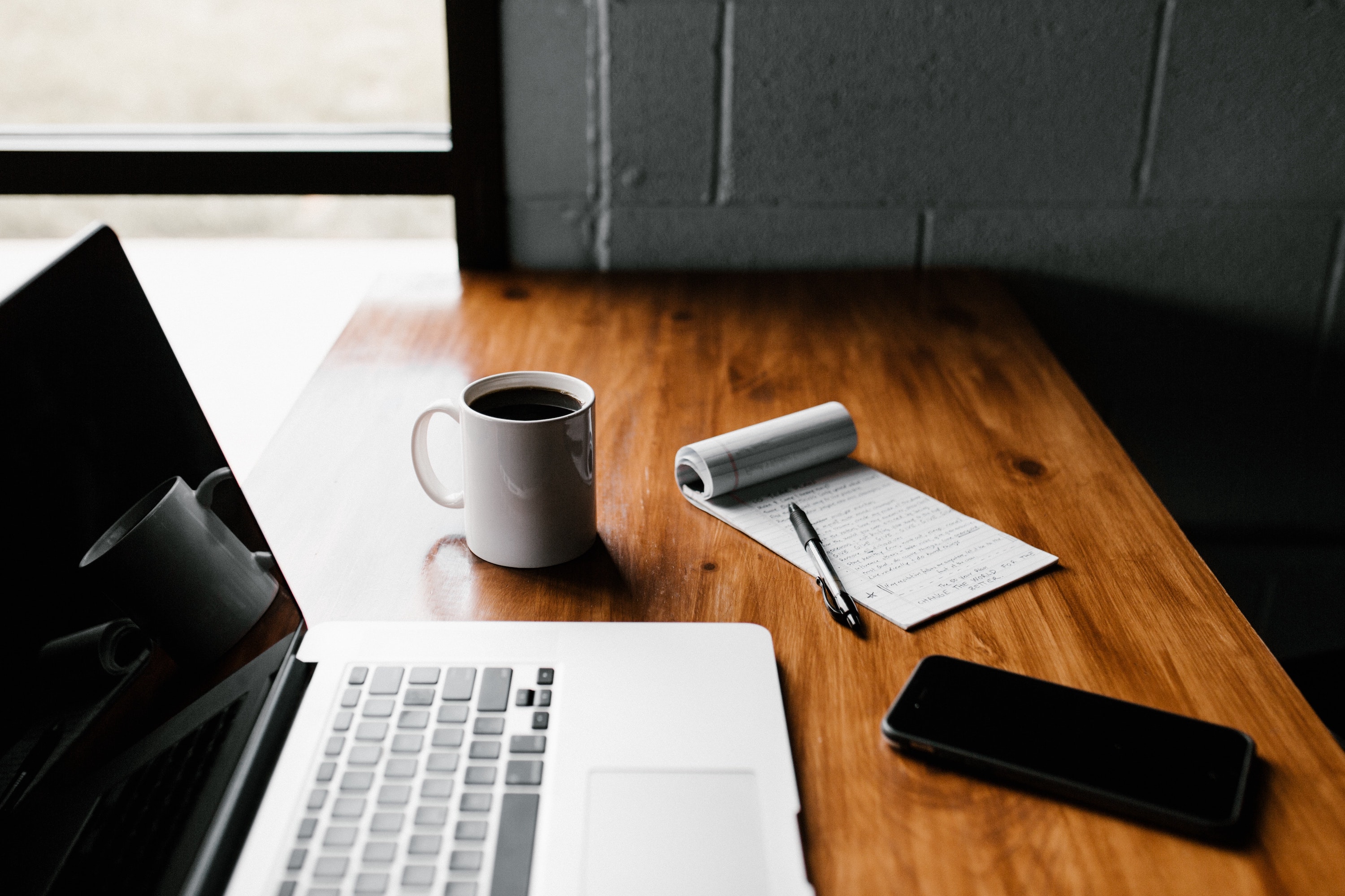 A laptop, a mug of coffee, a notepad with a pen, and a phone on a wooden desk
