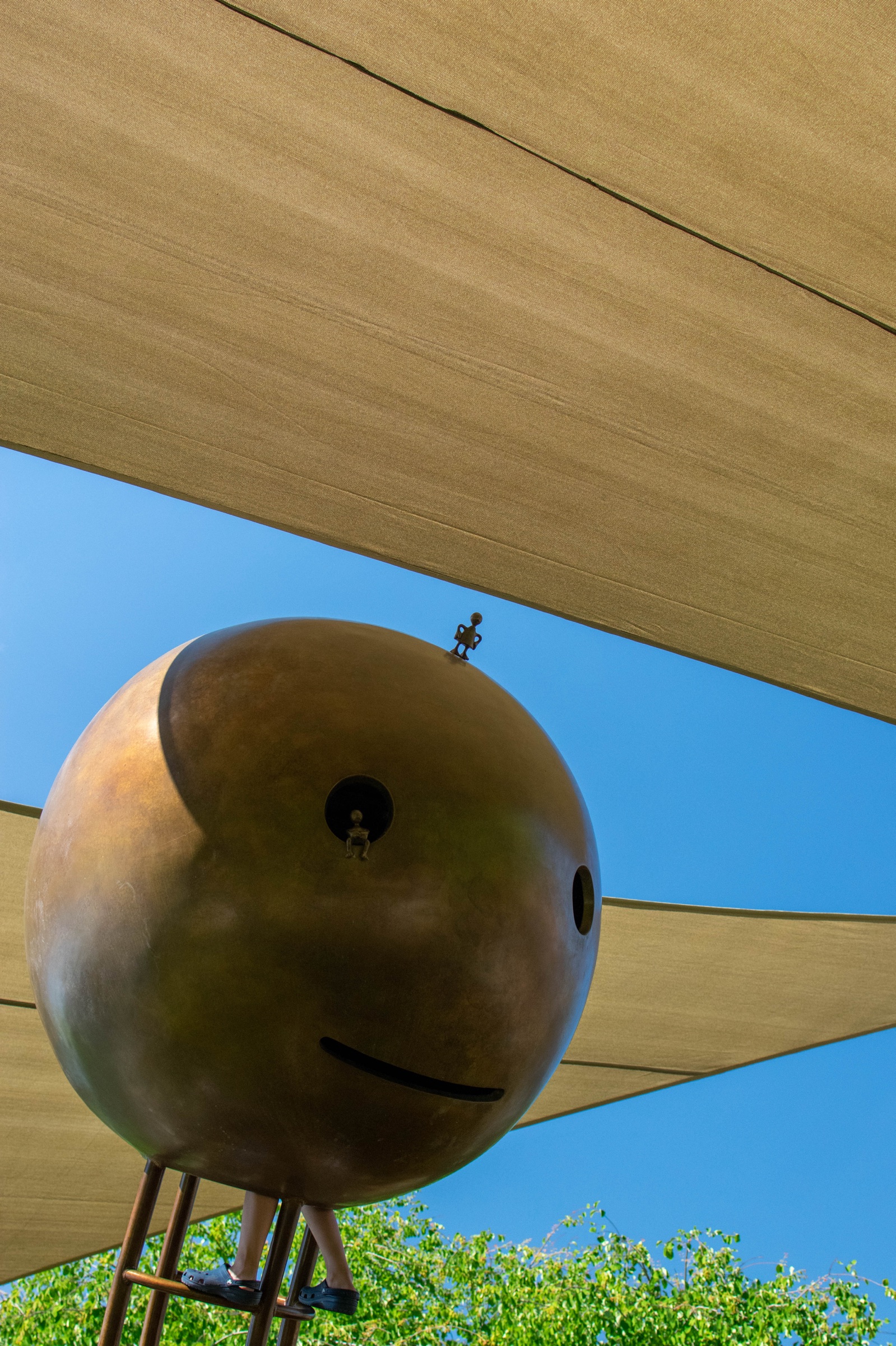 A bronze sculpture of a round smiling head on thin legs beneath tan shade sails and a blue sky