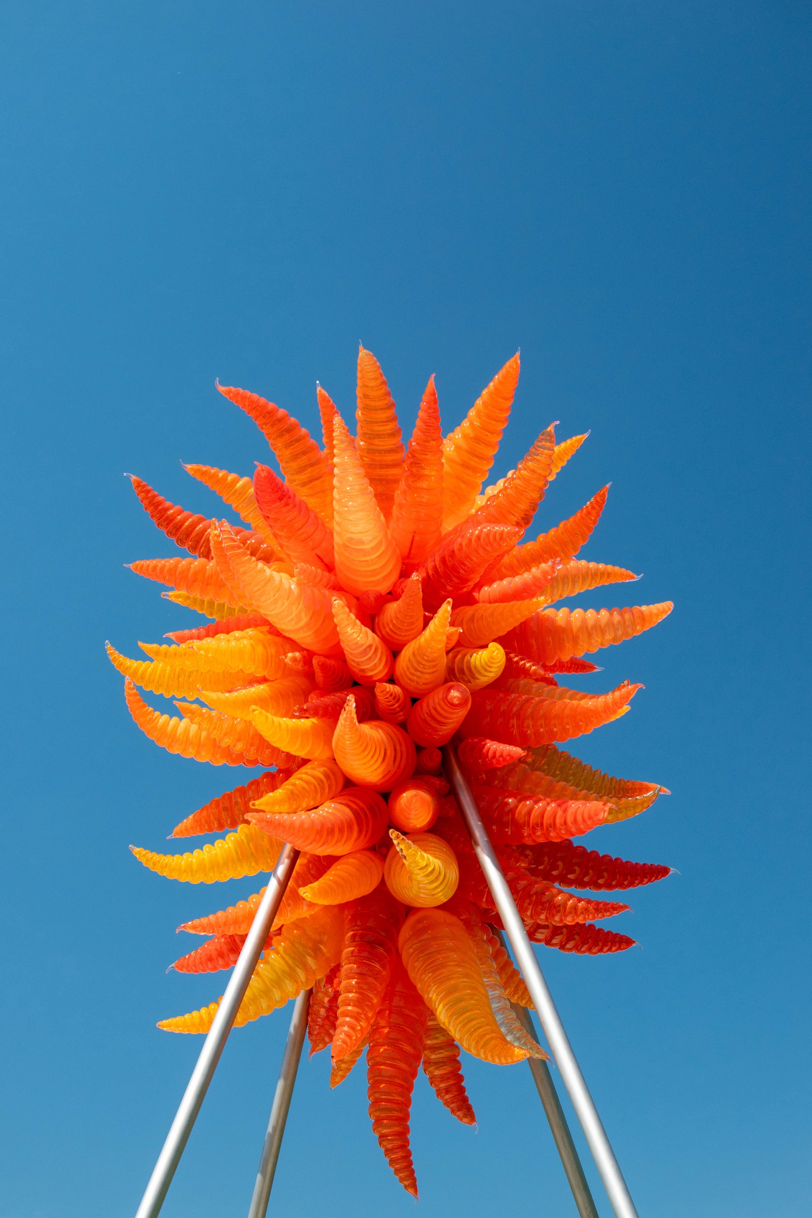 A spiky orange and red blown glass sculpture on metal legs against a clear blue sky