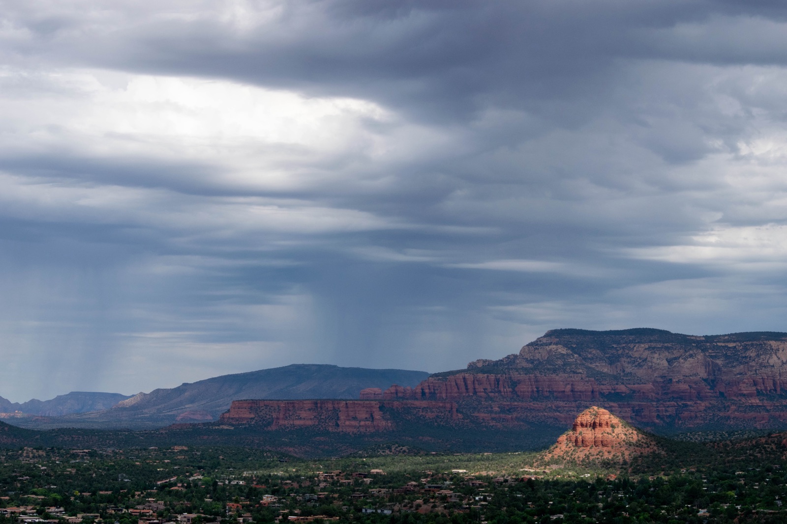 Rain falling from storm clouds over distant mesas, with a sunlit red rock butte below